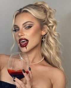 Brookelle McKenzie com corset preto, colar de pérola, cabelo loiro meio preso, taça de "vinho" e maquiagem de vampira