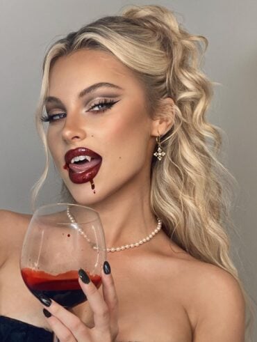 Brookelle McKenzie com corset preto, colar de pérola, cabelo loiro meio preso, taça de "vinho" e maquiagem de vampira