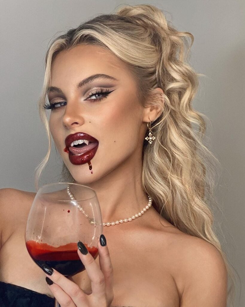 Brookelle McKenzie com corset preto, colar de pérola, cabelo loiro meio preso, taça de "vinho" e maquiagem de vampira