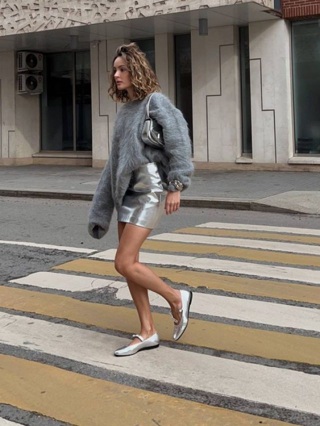 Mulher loira de cabelo curto e ondulado, usando tricô de pelinhos em cor cinza, saia metalizada e sapato boneca metalizado.