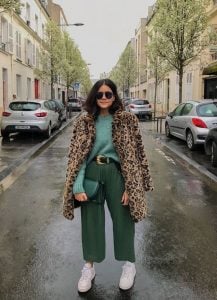 Mulher, no meio da rua, vestindo uma blusa verde claro, uma calça verde escuro, uma bolsa em outro tom de verde e um casaco ted de oncinha