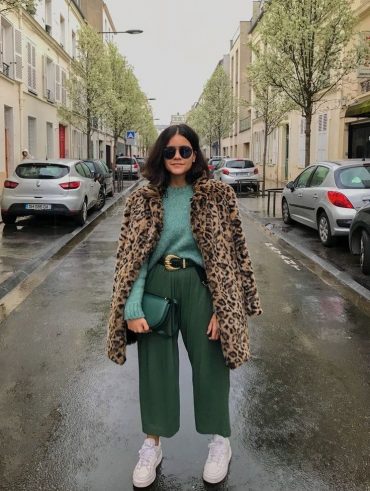 Mulher, no meio da rua, vestindo uma blusa verde claro, uma calça verde escuro, uma bolsa em outro tom de verde e um casaco ted de oncinha
