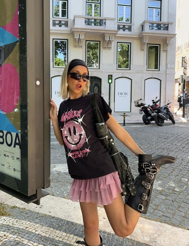 Menina branca se apoiando em uma placa, vestindo uma blusa preta e rosa com uma minissaia rosa
