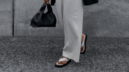 Pernas de uma mulher de calça, segurando uma bolsa preta e com chinelo preto e branco