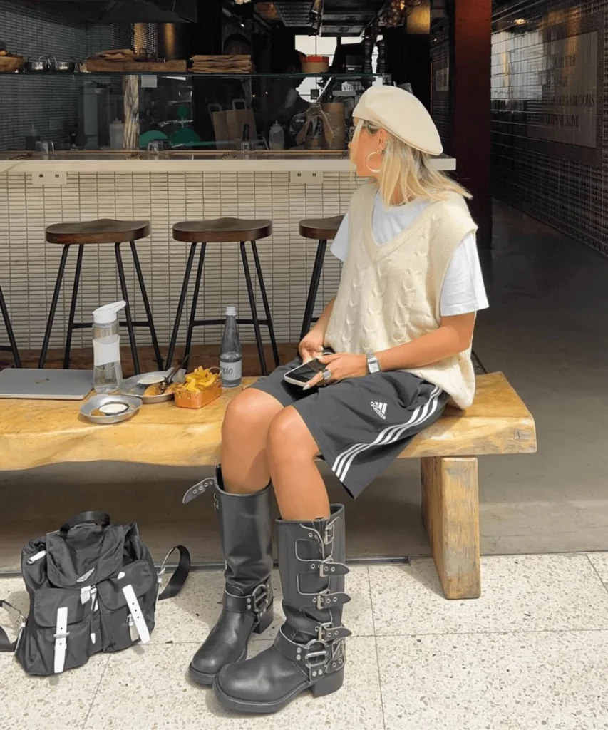 Mulher branca sentada em um banco vestindo bermuda, colete de tricô e biker boots