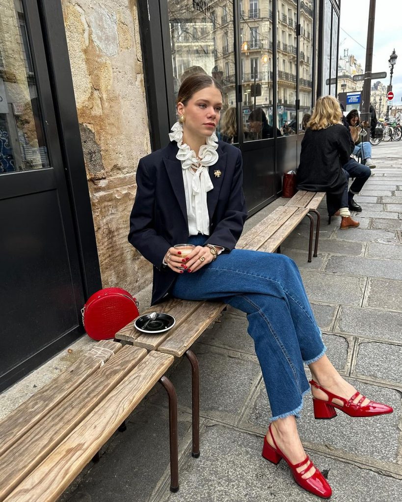 Mulher branca jovem sentada em banco de madeira. Ela usa blazer azul noite, calça jeans reta, sapato boneca vermelho