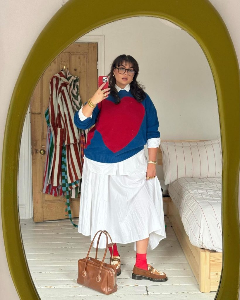 Francesca Perks tirando foto em frente ao espelho. Ela veste tricô sobreposto à camisa branca, saia midi branca, meia vermelha aparente e mocassim de camurça