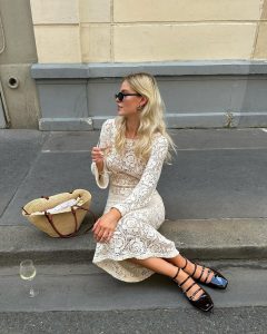 Mulher branca loira sentada em cordão de rua. Ela usa vestido feito à mão off-white, maxi bag de palha e calçado boneca preto