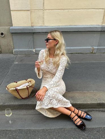 Mulher branca loira sentada em cordão de rua. Ela usa vestido feito à mão off-white, maxi bag de palha e calçado boneca preto