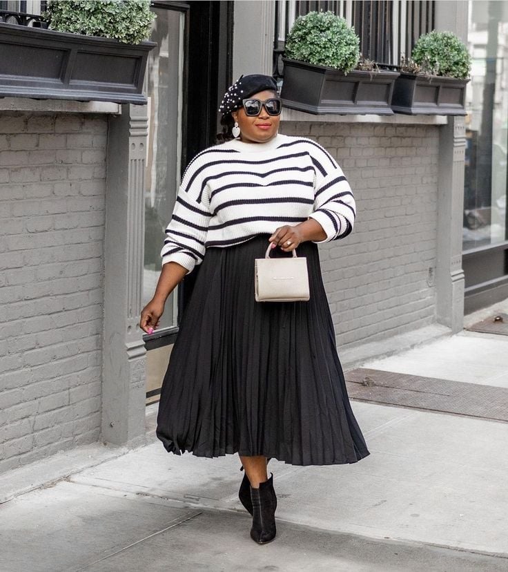 Mulher negra, plus size, com saia preta, bolsa branca e blusa de tricô listrado branco e preto