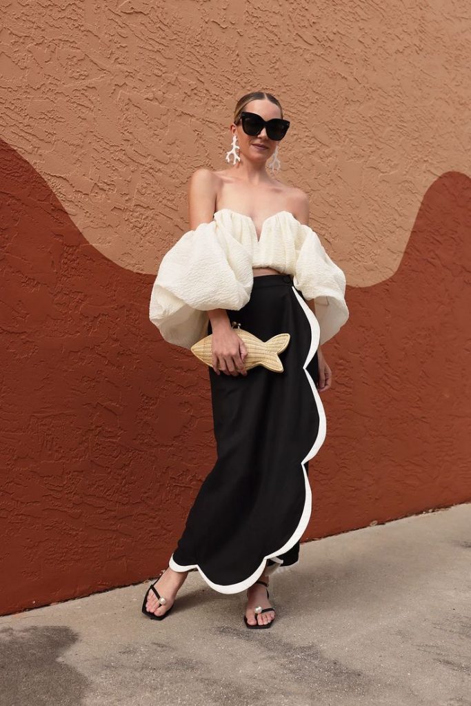 Mulher branca, com saia preta com detalhe branco, bolsa de peixe e blusa branca