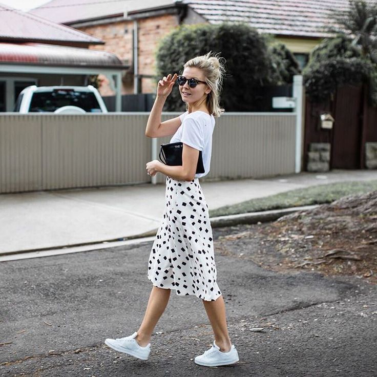 Mulher branca, com saia branca estampada, bolsa preta, blusa branca e tênis branco