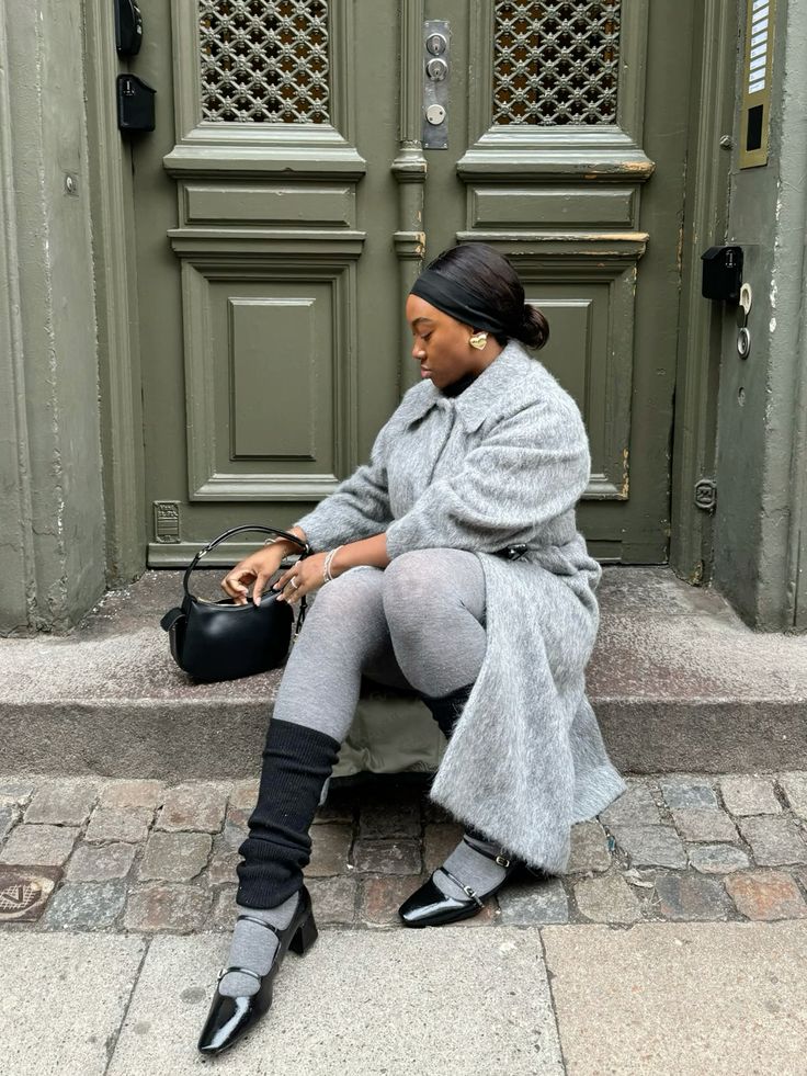 Mulher negra sentada em frente a uma porta. Ela usa faixa de cabelo, maxi brinco dourado, sobretudo cinza, meia-calça cinza, polaina preta, shoulder bag e calçado boneca preto