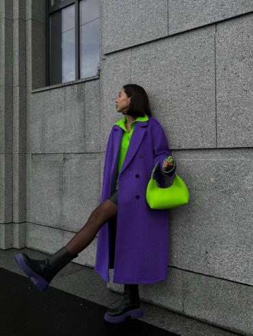 Mulher escorada na paredes, com a perna levemente levantada, usando um look verde , sobretudo roxo e bolsa verde