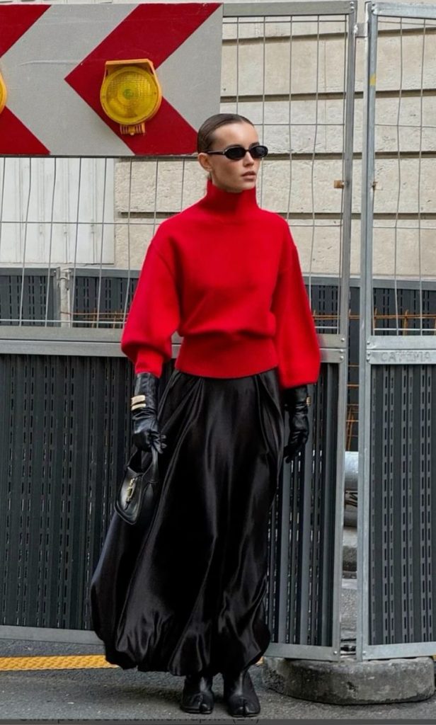 Mulher jovem usando óculos de sol, tricô vermelho de gola alta, luva, bolsa preta, saia de cetim ampla e calçado pata de vaca
