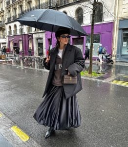 Mulher jovem segurando guarda-chuva. Ela veste blazer preto, moletom marrom, saia preta balonê e bota