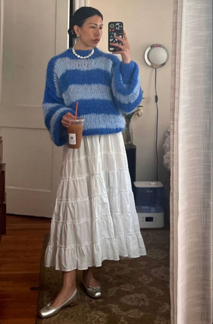 Mulher jovem tirando foto em frente ao espelho. Ela veste tricô azul listrado, maxi skirt branca de fibra natural e sapatilha metalizada