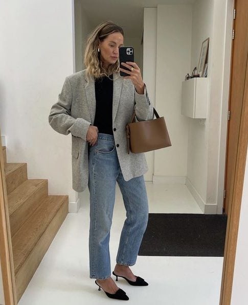 mulher vestindo blazer cinza e calça jeans clara com sapato scarpin. Ela está em frente ao espelho