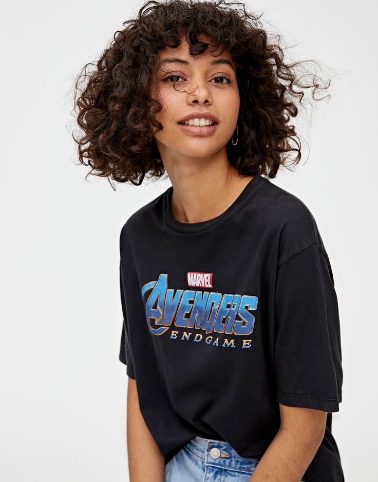 Mulher com cabelo cacheado, curto e reto, no estilo bob, com franja. Usa camiseta preta e calça jeans.