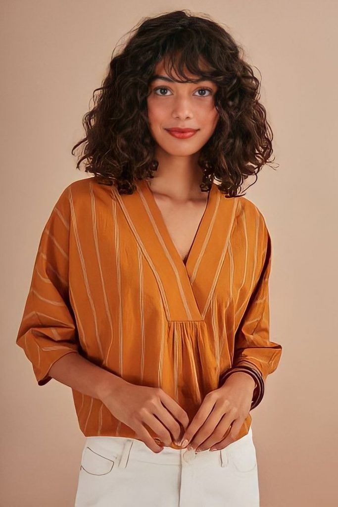 Mulher com cabelo cacheado e curto, no estilo bob, com franja. Usa blusa terracota e calça branca.
