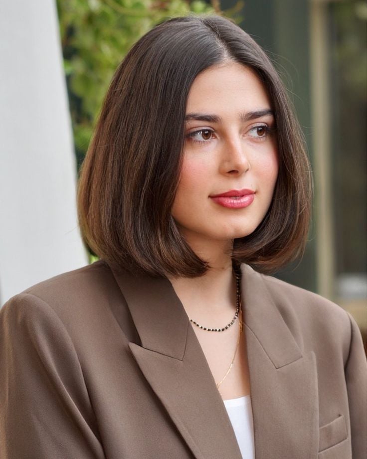 Mulher branca com blazer marrom, blusa branca, colar e cabelo castanho bubble bob