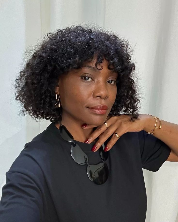 Mulher com cabelo cacheado e curto, no estilo bob, com franja. Usa blusa preta, brinco e colar grande e preto.