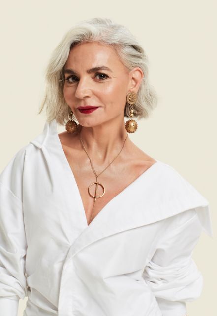 Mulher branca de 60 anos, usando maquiagem suave e cabelo curto, no estilo bob. Usa camisa branca e lisa com colar e brincos.