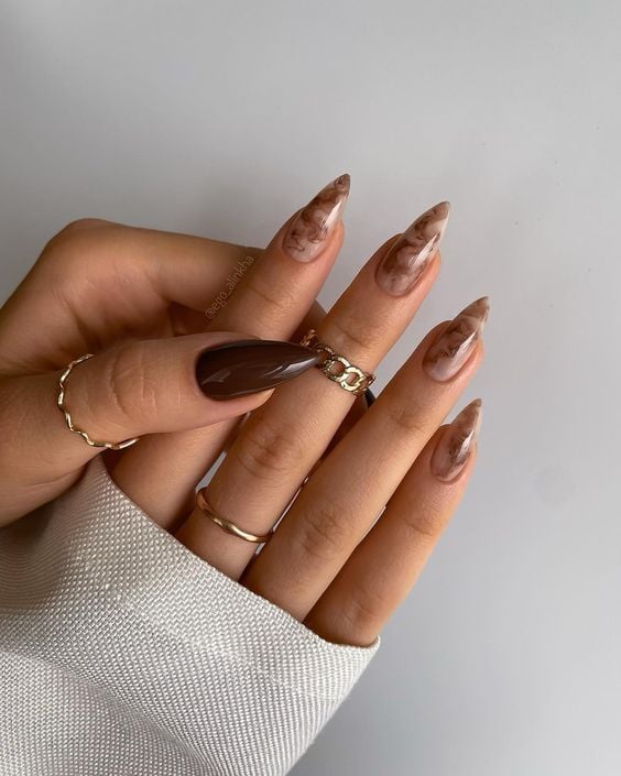 Mão com manga branca, anéis e mãos meio sobrepostas. Unha marrom com a estética coffee