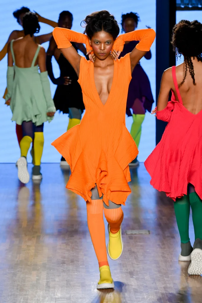 Mulher negra desfilando com sapato amarelo, meia laranja, vestido e mangas laranja 