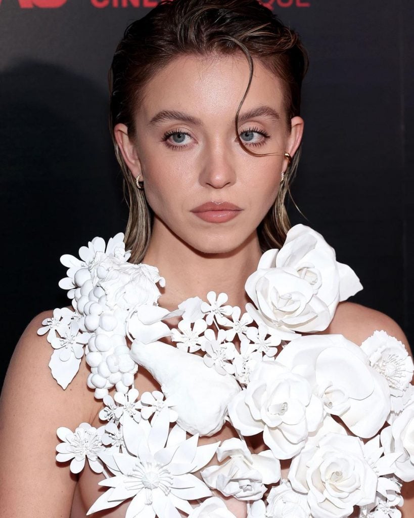 Sydney Sweeney com cabelo curto com franja, com penteado em gel. Usa vestido com aplicações de flores