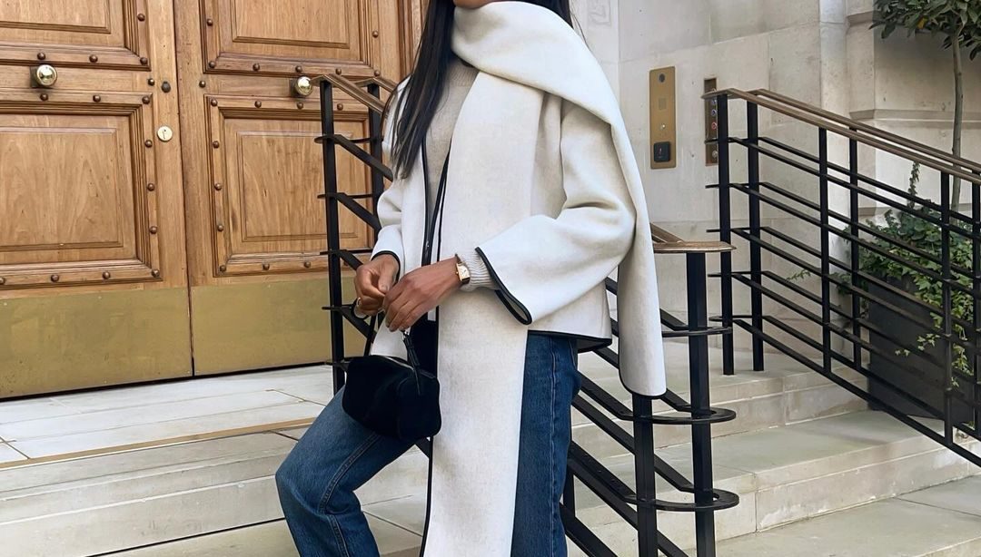 Mulher não branca. Ela usa cachecol off-white, casaco off-white, calça jeans reta e loafer branco