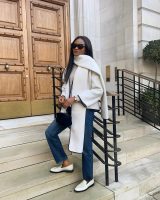 Mulher não branca. Ela usa cachecol off-white, casaco off-white, calça jeans reta e loafer branco