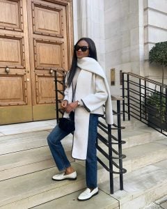 Mulher não branca. Ela usa cachecol off-white, casaco off-white, calça jeans reta e loafer branco