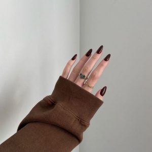 Mão branca com manga marrom, anéis e unha marrom quente