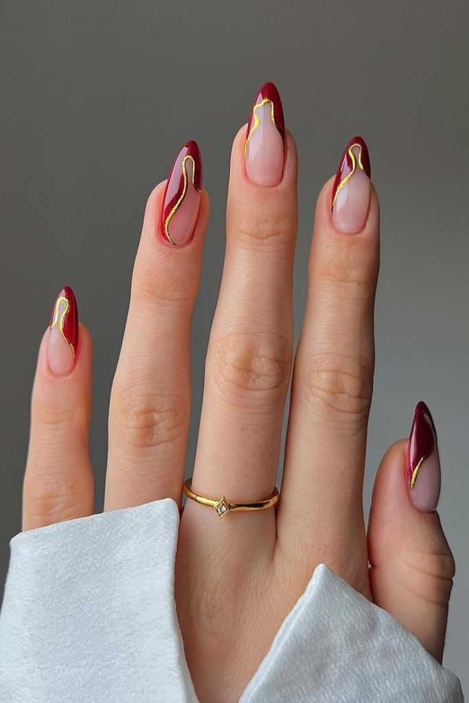 Mão branca com manga branca e anel, a unha está com desenhos orgânicos na cor vermelho cereja e dourado