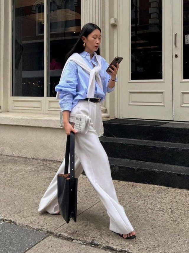 Mulher não branca jovem. Ela usa camisa social azul listrado oversized, tricô off-white e calça pantalona de alfaiataria