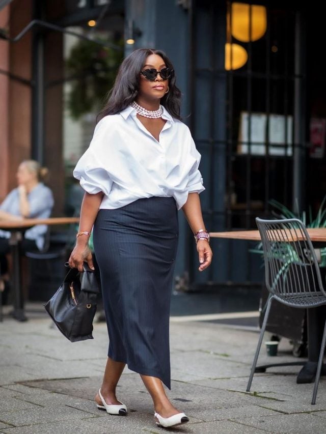 Mulher negra. Ela usa colar de pérolas, camisa social branca, saia midi risca de giz azul escuro, bolsa preta e slingback bicolor