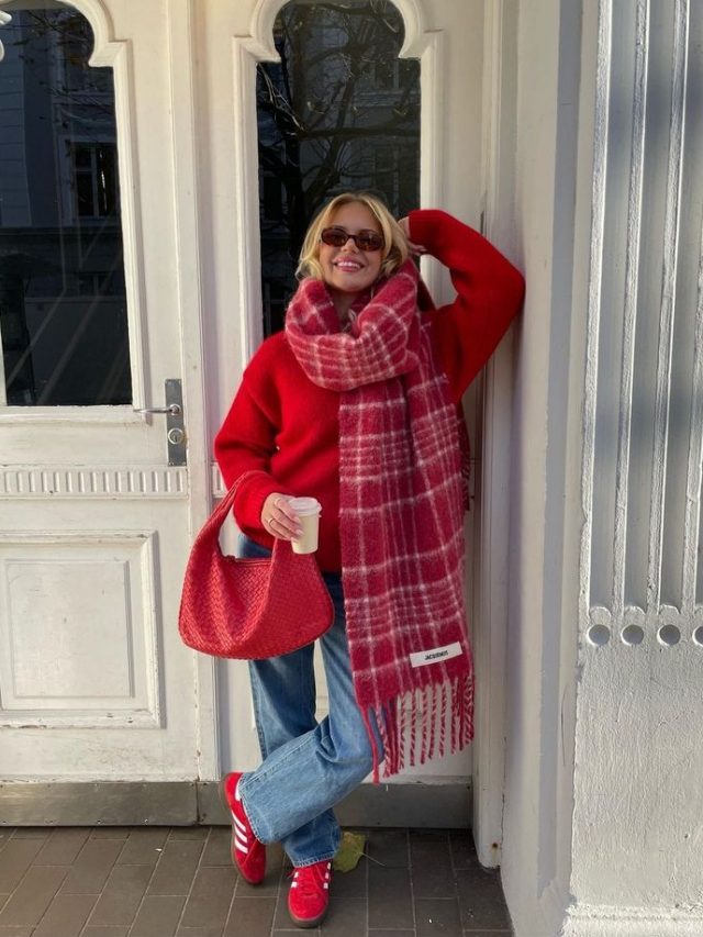 Mulher apoiada em um viga, com sapato, bolsa, blusa e cachecol vermelho. Além de usar calça jeans
