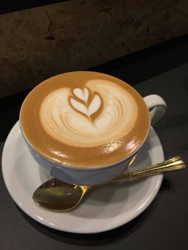 Imagem com uma xícara de café em cima de um pires. O café é um capuccino com desenho de uma folha.