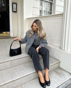 Mulher jovem sentada em escadaria. Ela veste casaco cinza, blusa cinza, calça jeans de lavagem escura e sapatilha básica preta envernizada