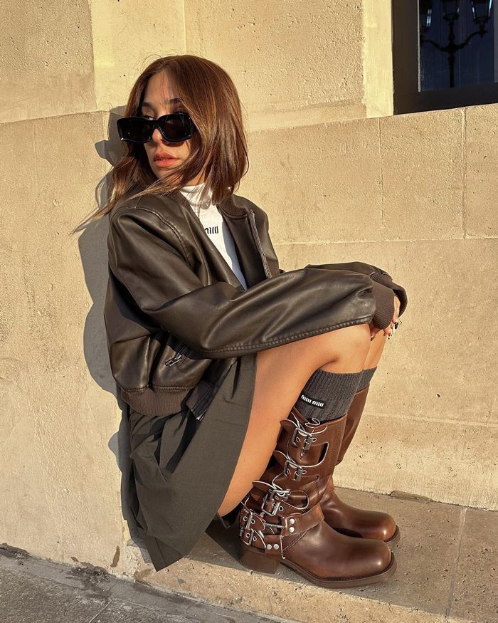 Mulher jovem branca sentada. Ela usa óculos de sol preto, jaqueta de couro, blusa Miu Miu branca, minissaia de alfaiataria cinza, meia três quartos cinza Miu Miu e bota biker marrom Miu Miu