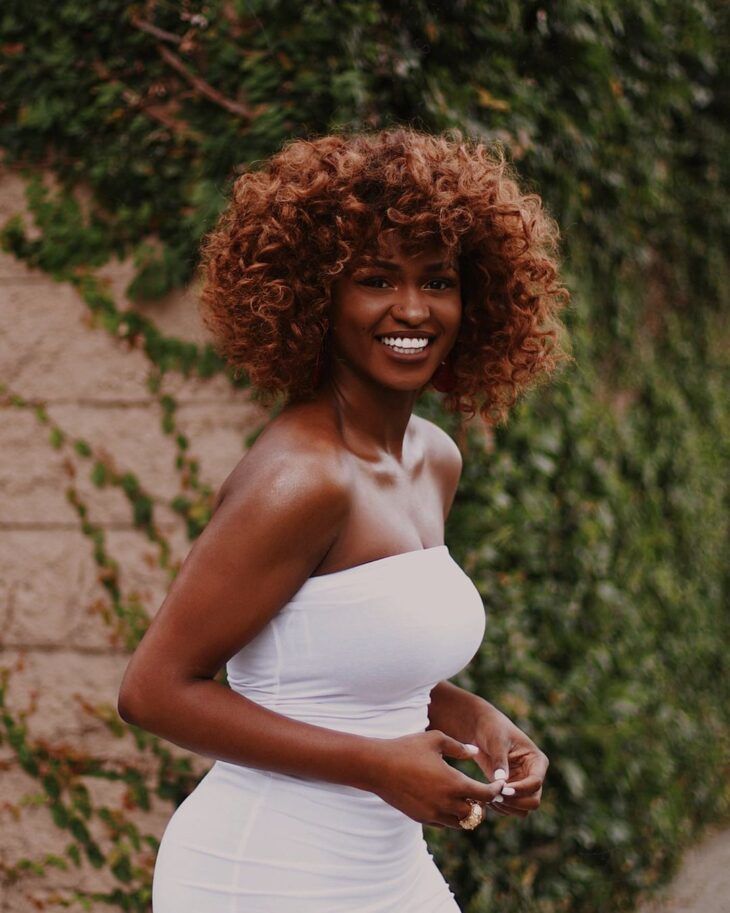 Mulher negra com vestido branco e cabelo crespo ruivo nude