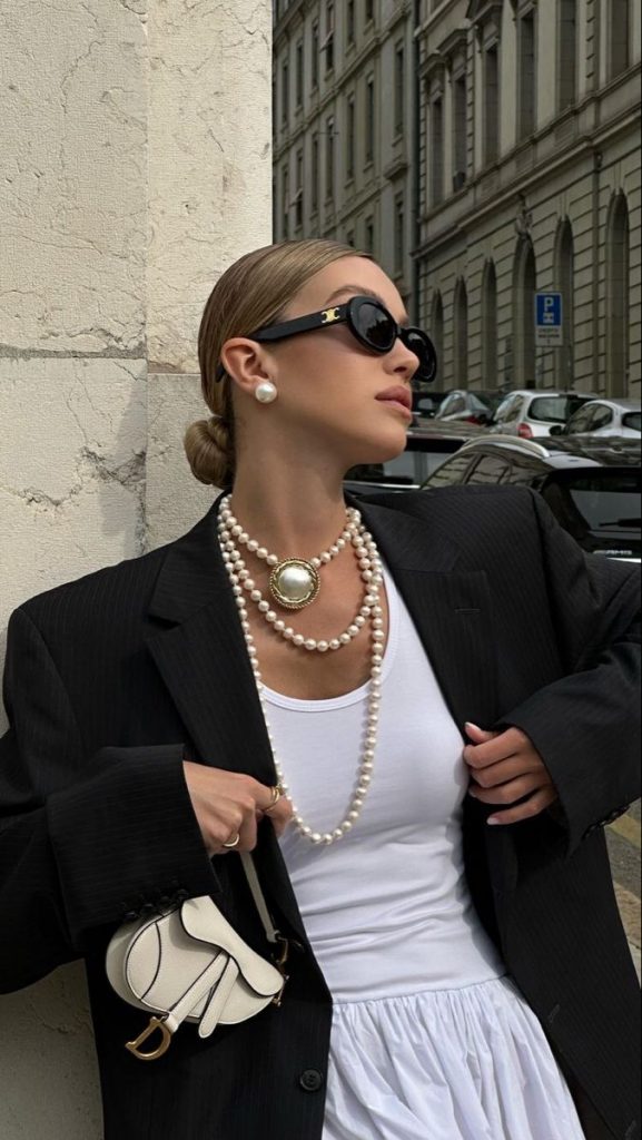 Mulher branca com blusa branca, blazer preto e mix de colares de pérola