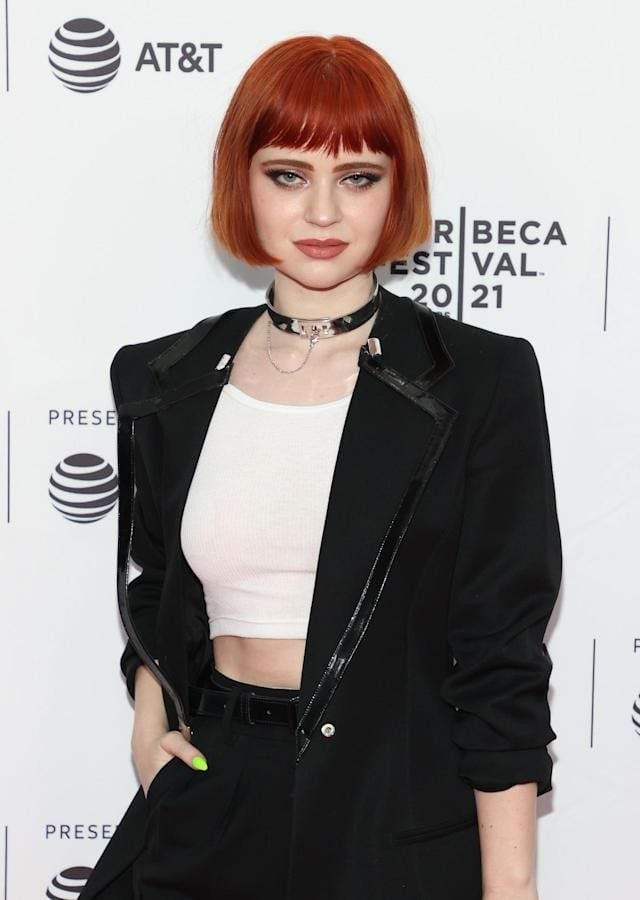 Mulher branca com blusa branca, casaco e calça preta e cabelo curto ruivo bob