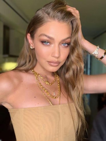 Gigi Hadid com blusa tomara que caia, acessórios cabelo loiro ondulado e maquiagem neutra