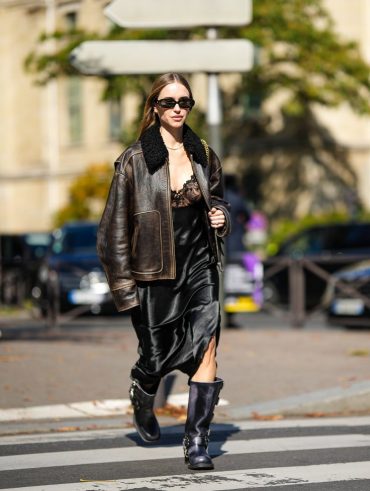 Mulher branca usando óculos de sol. Ela usa jaqueta estonada, slip dress e bota biker