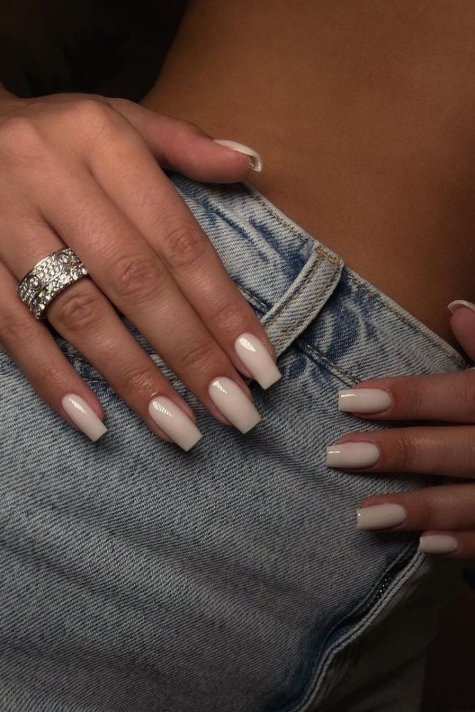 Mão branca com anel, apoiadas em uma calça jeans, com unha bailarina branca