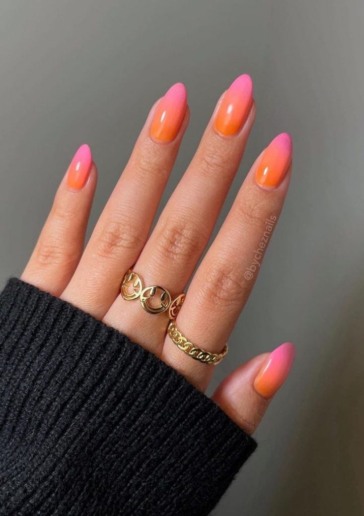 Mão branca com manga preta, anéis e unha degradê rosa e laranja