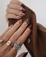 Mãos brancas com anéis apoiadas com a unha marrom degradê com design de meia-lua