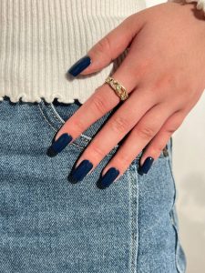Mão branca com anel apoiada na calça com a unha quadrada azul marinho